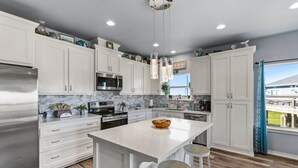 A modern kitchen with white cabinets, a large island, stainless steel appliances, and a view of a residential area. - Steps to the Beach l Modern Interior (Crystal Beach)