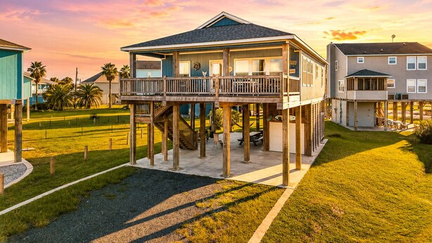 A two-story house with a balcony, elevated on stilts, surrounded by a grassy yard and palm trees. - Steps to the Beach l Modern Interior (Crystal Beach)