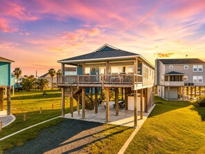 A two-story house with a balcony, elevated on stilts, surrounded by a grassy yard and palm trees.