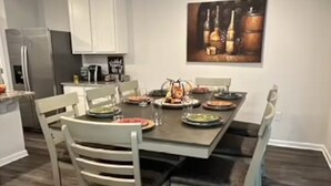 A dining area with a wooden table set for four, surrounded by white chairs. A painting of bottles hangs on the wall above the table. - Welcoming Spacious Crestview Home w/ Pool Access & Fire Pit (Crestview)