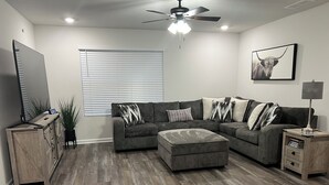 A living room with a sectional sofa, ottoman, ceiling fan, and a mounted TV. - Welcoming Spacious Crestview Home w/ Pool Access & Fire Pit (Crestview)