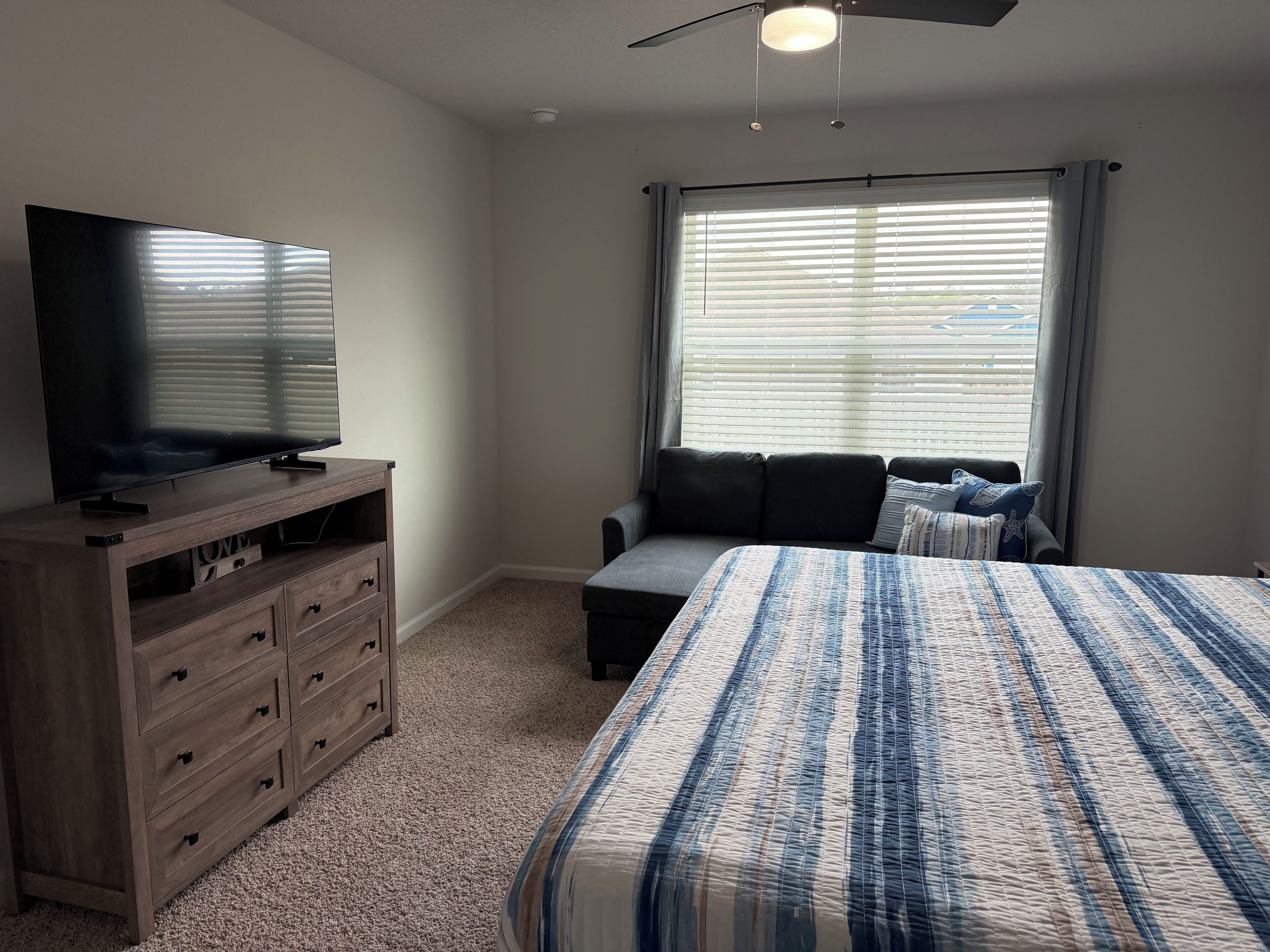 A bedroom with a bed, a TV on a wooden dresser, a window with blinds, and a ceiling fan.