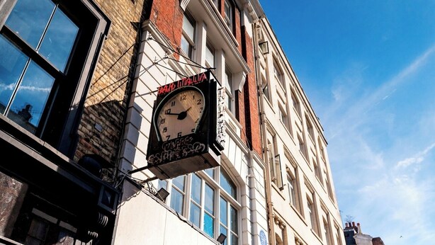Une horloge traditionnelle, ornée des inscriptions « BAR ITALIA » et « ESPRESSO », est fixée à un bâtiment doté de grandes fenêtres et d’une façade en briques.