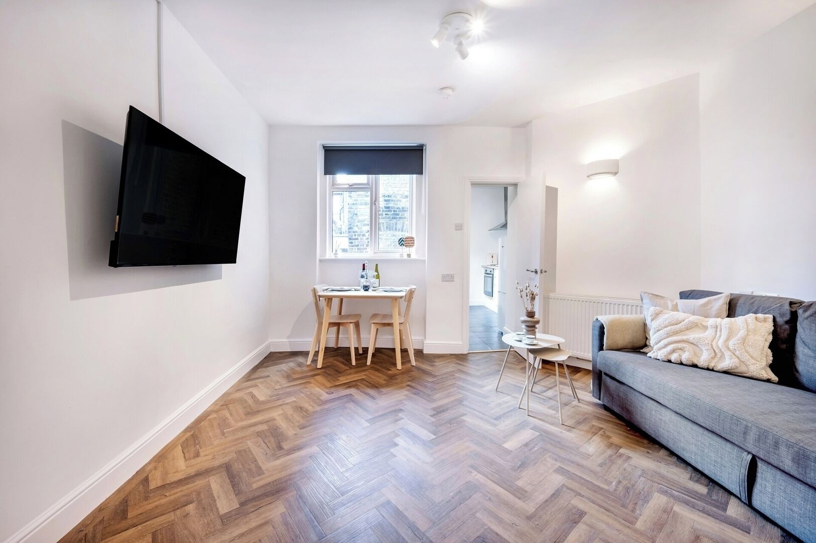 Un salon moderne avec un parquet en bois disposé en chevrons, un canapé gris agrémenté de coussins décoratifs, un téléviseur à écran plat fixé au mur et un coin repas avec une petite table ronde et des chaises.