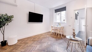 Un salon moderne doté d’un téléviseur à écran plat, d’un coin repas avec une table et des chaises, d’une plante en pot et d’une vue sur l’extérieur par une fenêtre.