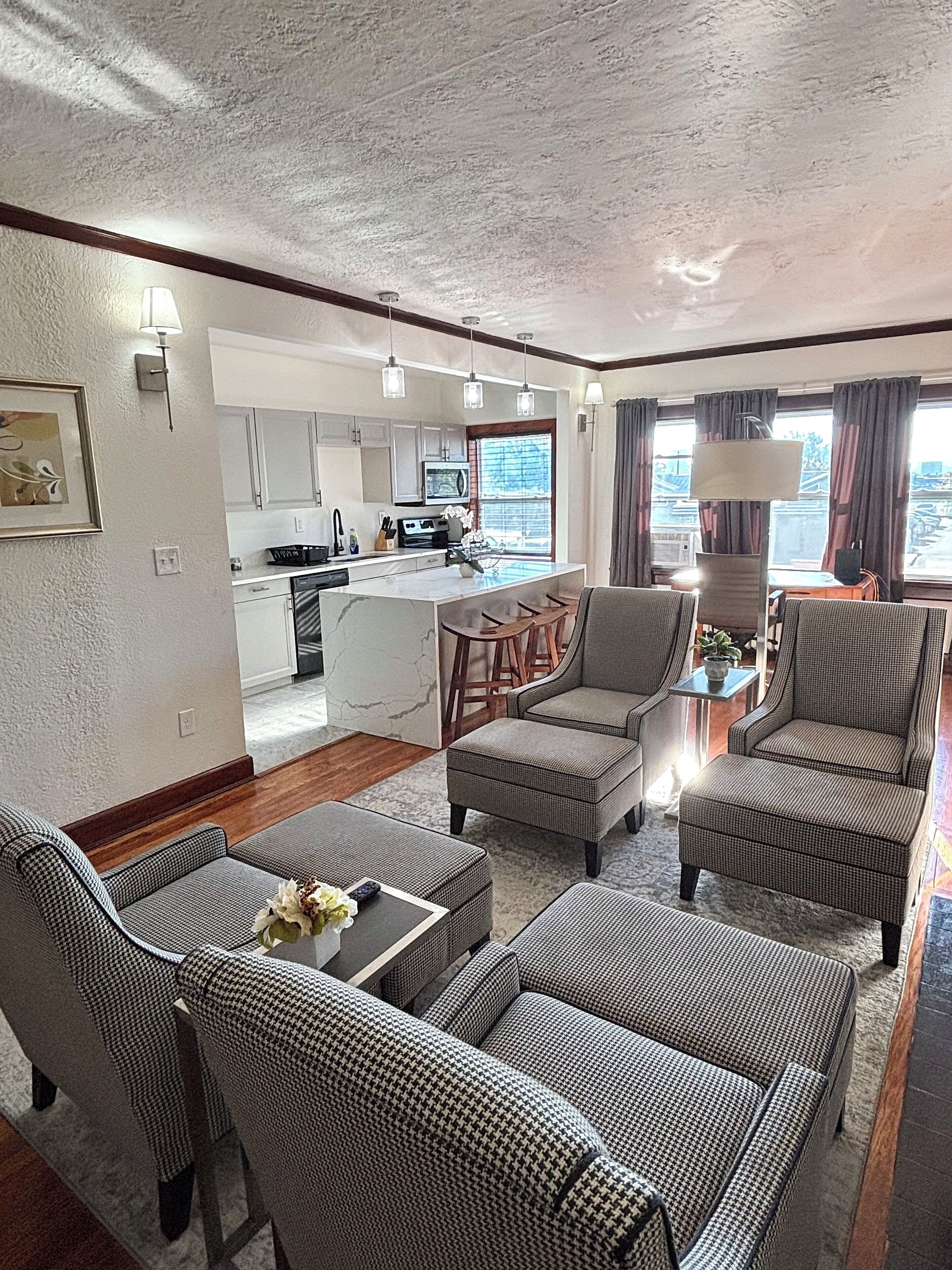 A spacious living area with a kitchen in the background, featuring a dining table, sofas, and a large window with curtains.