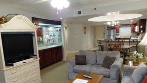 Ein Wohnzimmer mit grauem Sofa, einem Holztisch mit Papieren, einem Fernseher auf einem weißen Schrank und einer Küche im Hintergrund.