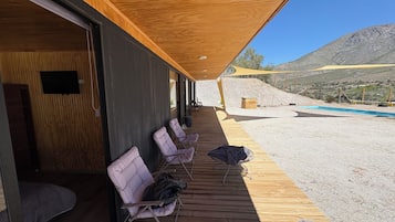 A covered outdoor seating area with wooden flooring and chairs, overlooking a pool and a mountainous landscape.