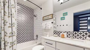 Une salle de bain avec une baignoire blanche, des toilettes et un lavabo avec un miroir au-dessus. Des serviettes sont suspendues à un porte-serviettes et il y a un rideau de douche à motifs géométriques.