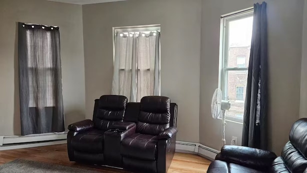 A living room with a dark leather sofa, a white fan, and a window with curtains. - Lovely fully equipped 4-bedroom apartment in charming Boston with parking. (Boston)