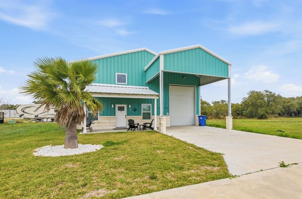 A two-story building with a green exterior and a white garage door. There is a palm tree in the front yard and a picnic table with chairs.