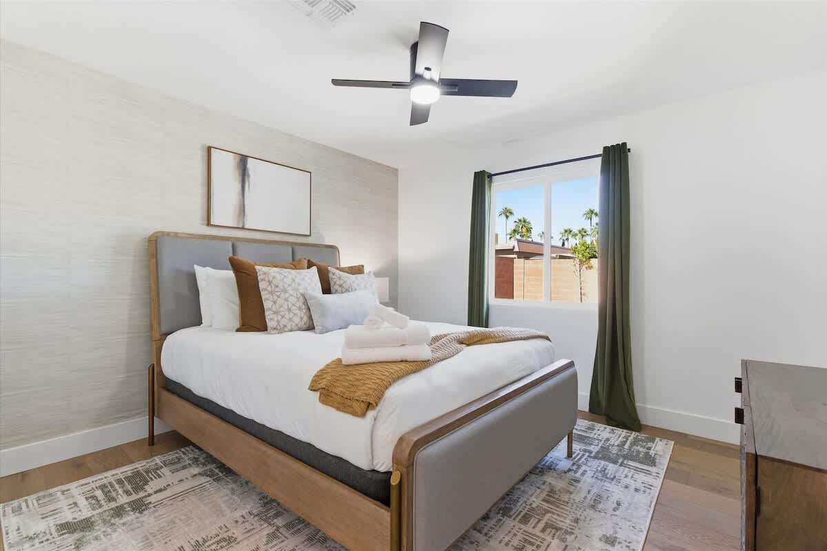 Une chambre à coucher avec un lit, un ventilateur de plafond, une fenêtre avec des rideaux et une vue sur des palmiers.