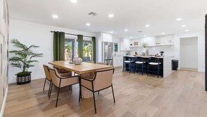 Uma cozinha moderna com armários brancos, uma área de refeições com mesa e cadeiras de madeira, e uma planta em vaso.