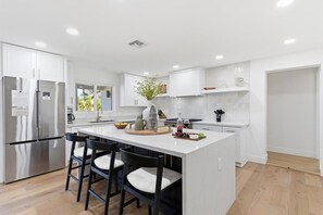 A modern kitchen with white cabinetry, a stainless steel refrigerator, a central island with bar stools, and a view of the outdoors.