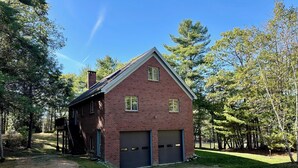 A two-story brick house with a garage, surrounded by trees and a dirt driveway.
