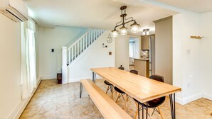 Un comedor amplio con una larga mesa de madera y bancos, una escalera que sube a otro nivel, y una cocina visible a través de un vano abierto.