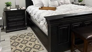 Quarto com estrutura de cama em madeira escura, roupa de cama branca, tapete estampado e arte decorativa nas paredes.