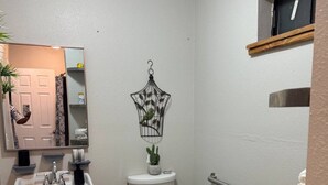 Uma casa de banho compacta com lavatório, sanita e toucador brancos. Uma gaiola decorativa pende na parede e há uma pequena janela sobre a sanita.