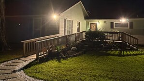 Uma casa com deck de madeira, caminho de pedra e gramado bem cuidado à noite.