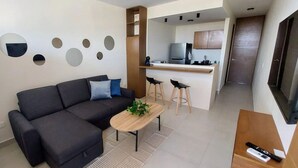 Een moderne woonkamer met een bank, salontafel en een keukenhoek met een koelkast en barstoelen.