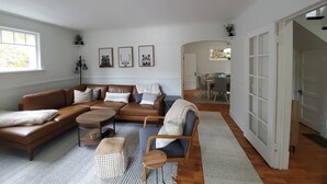 A spacious living room with a brown leather sofa, a coffee table, and a patterned rug. There are framed pictures on the wall and a dining area in the background. - Snowhawk House in Historic Wallace located close to many outdoor adventures! (Wallace)