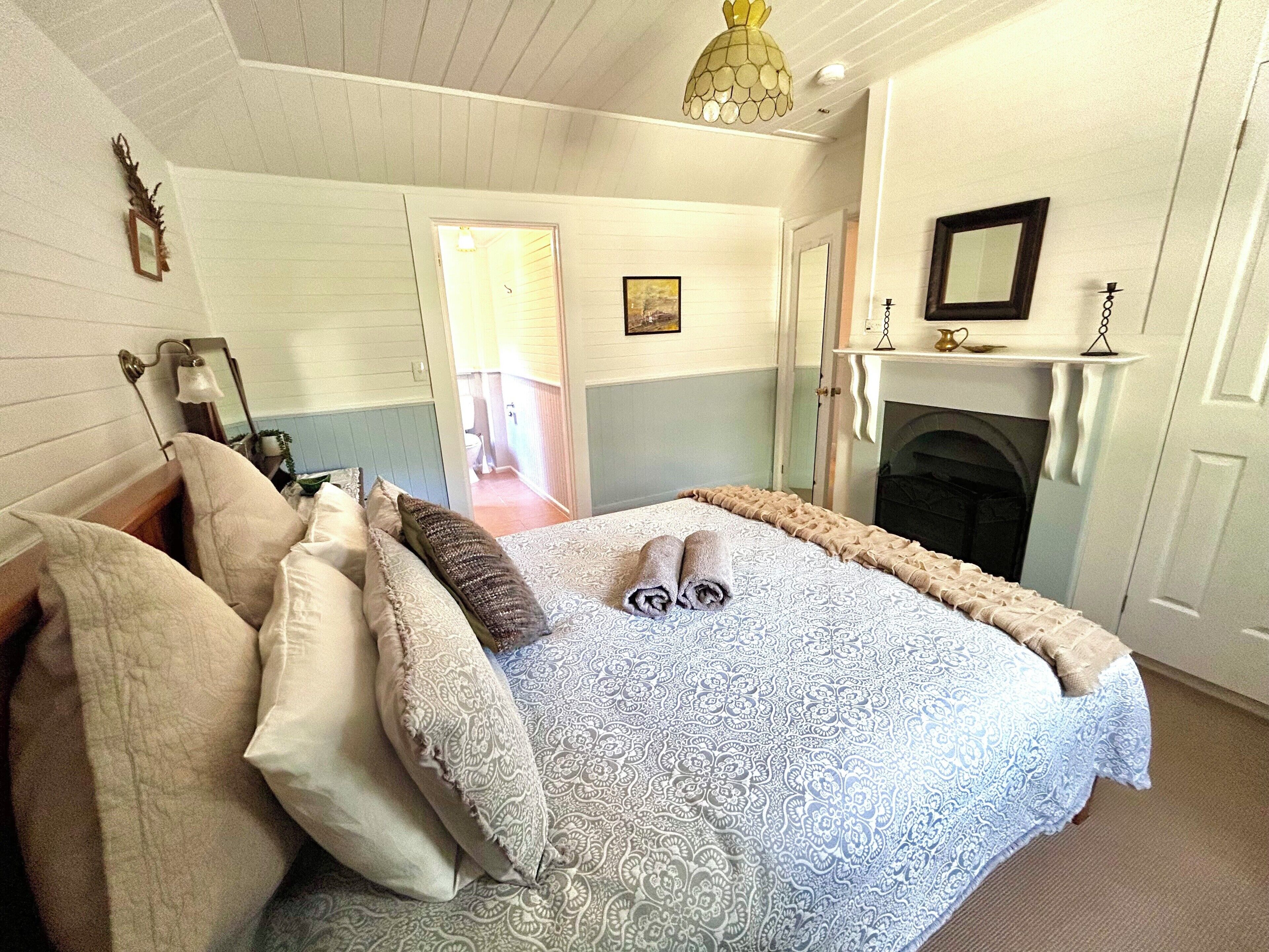 A bedroom with a bed, pillows, a mirror, a fireplace, and a door leading to another room.