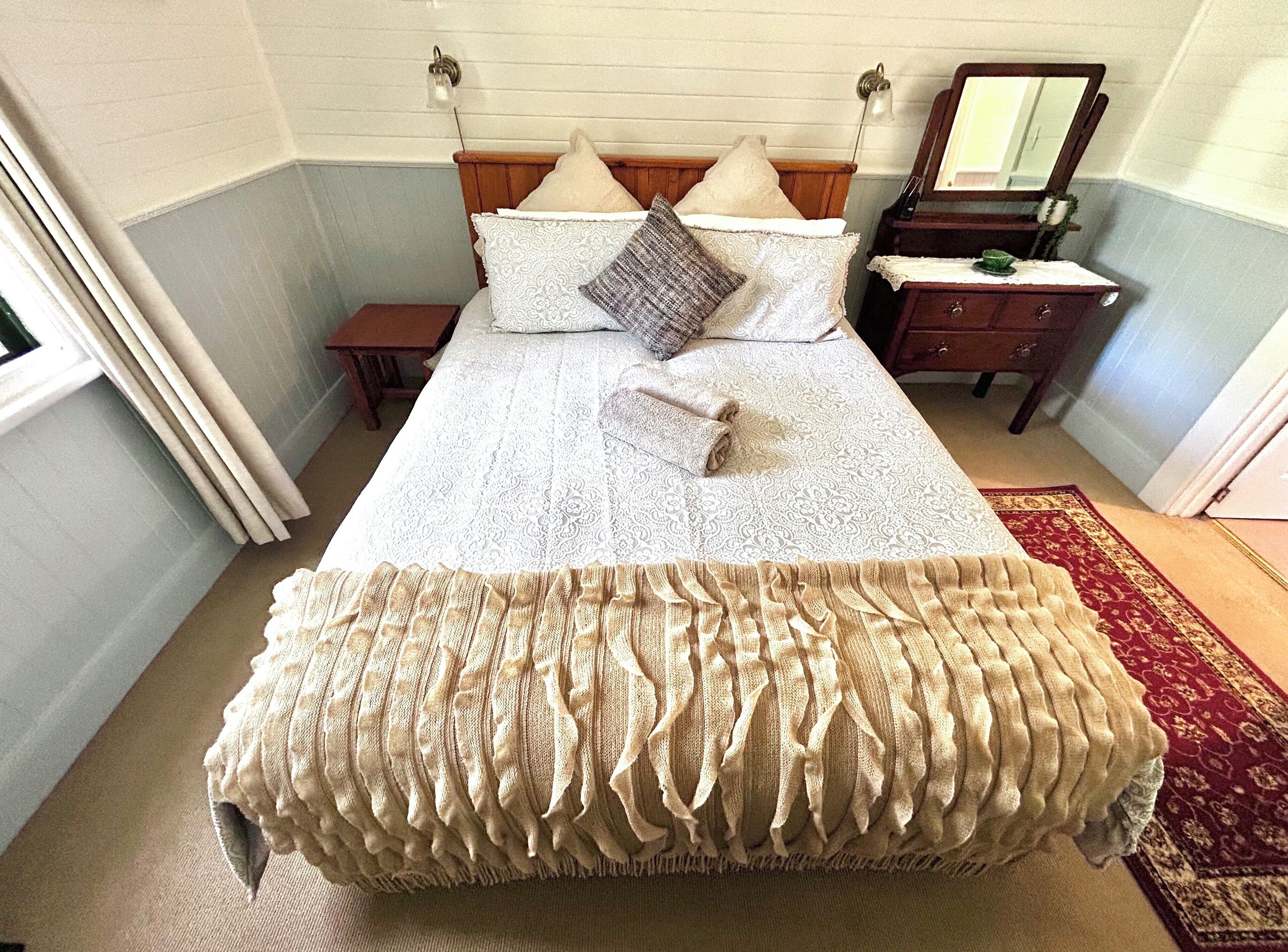 A neatly made bed with a patterned bedspread and pillows, a wooden headboard, a nightstand with a lamp, and a dresser with a mirror.