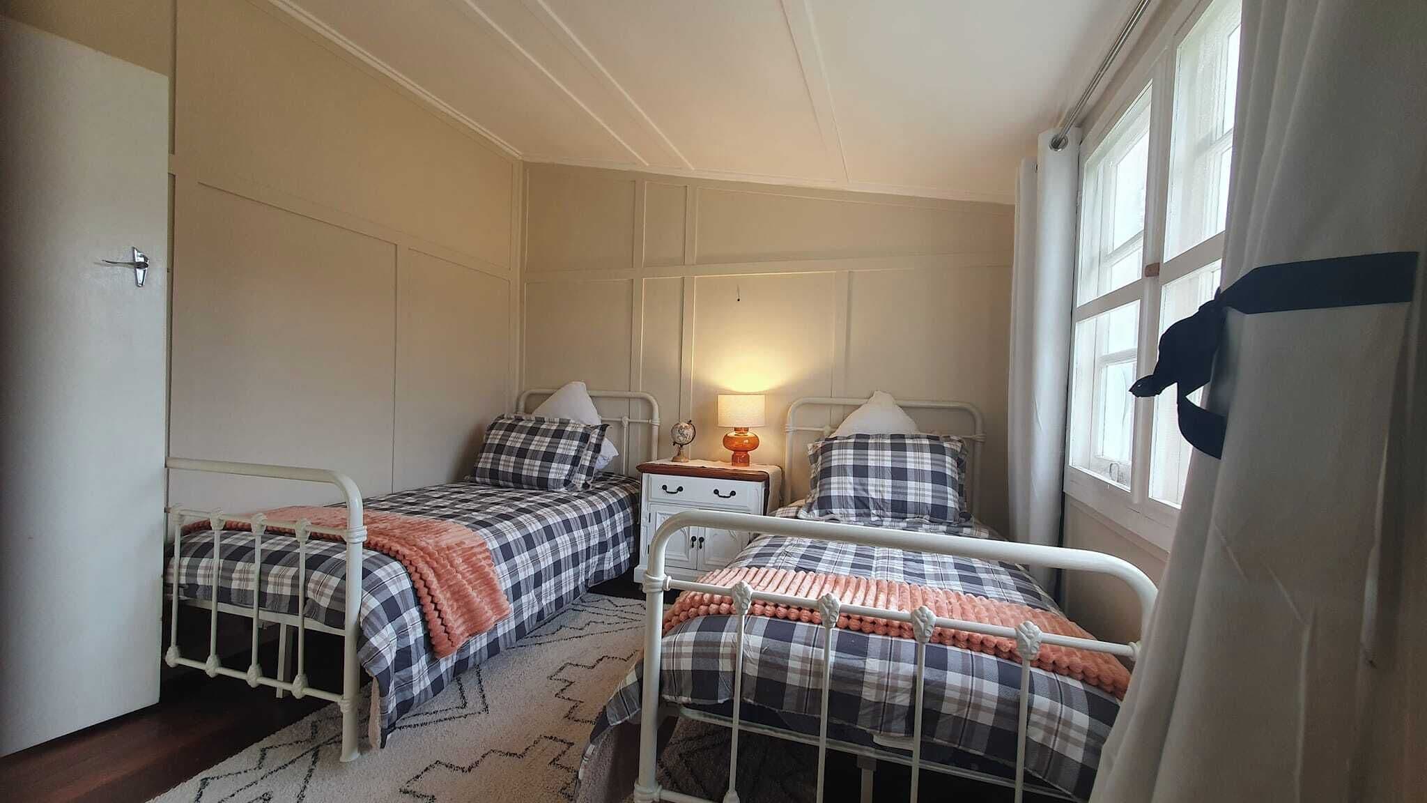 A small, cozy bedroom with two single beds, a nightstand with a lamp, and a window with curtains.