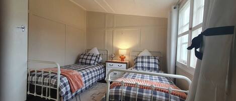 A small, cozy bedroom with two single beds, a nightstand with a lamp, and a window with curtains.