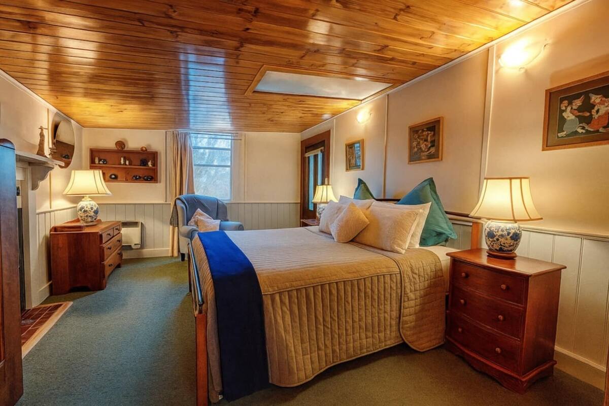 Un dormitorio con techo de madera, una cama con cubrecama azul y beige, dos mesitas de noche con lámparas, una silla, un aparador y una ventana con cortinas.