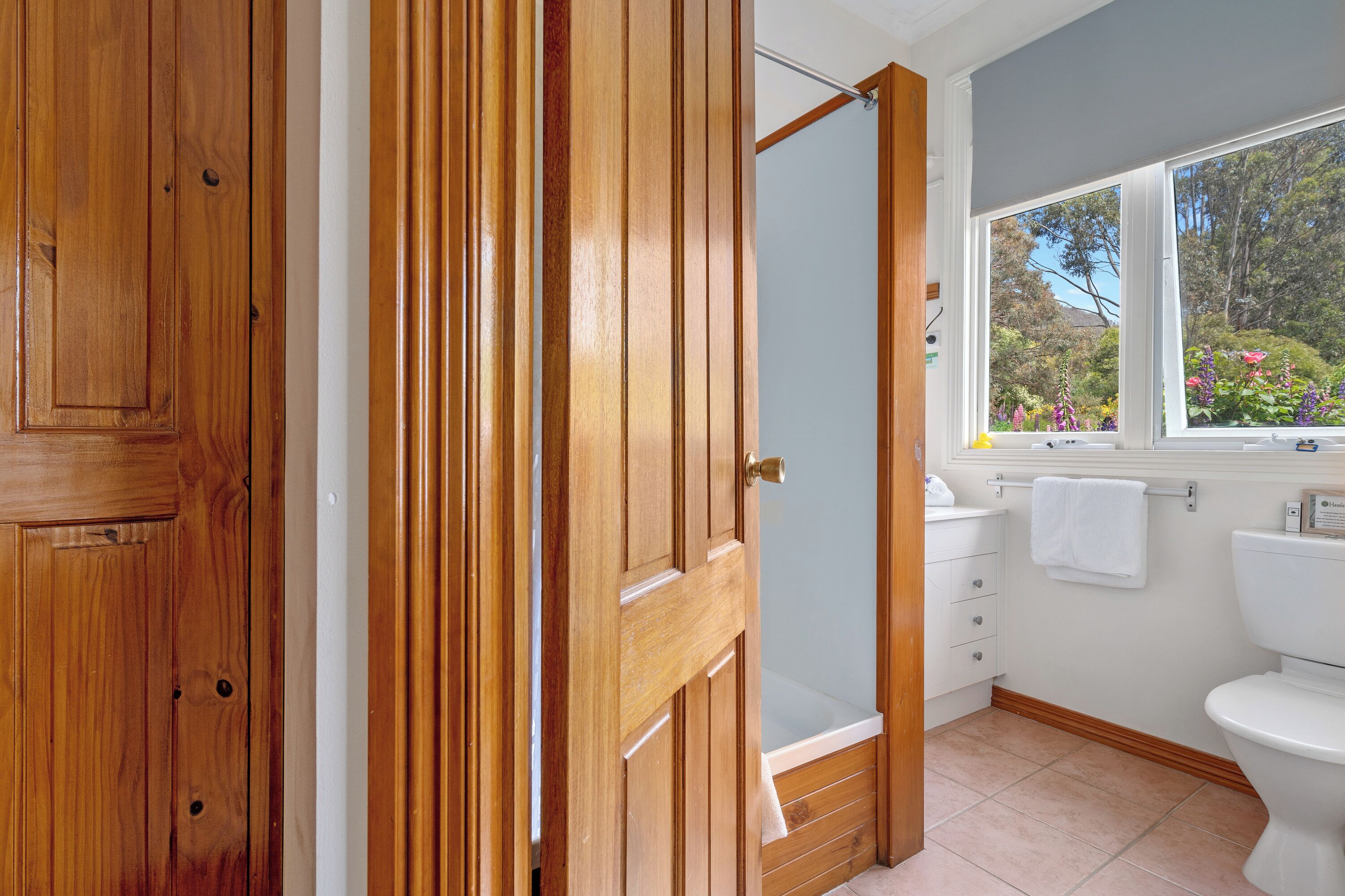 Une salle de bain avec une porte en bois, des toilettes blanches, une fenêtre donnant sur un espace verdoyant et un lavabo blanc.