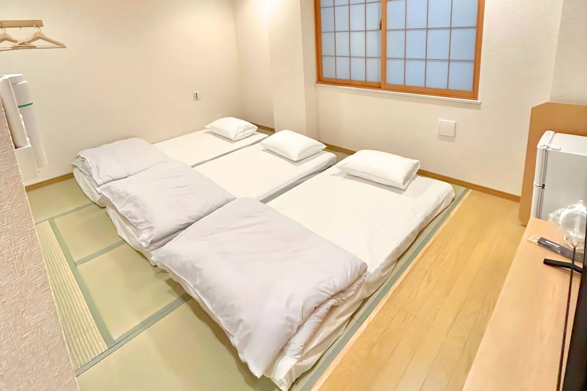 A hotel room with three beds, a wooden floor, a window with a grid pattern, and a small table with a bottle and a container.