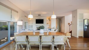 A modern kitchen with a wooden dining table, four wicker chairs, pendant lights, and a built-in TV. - Casa Piscina, Coogee Isyd (Coogee)