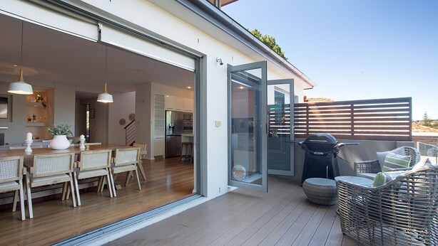 A modern kitchen with white cabinets and stainless steel appliances, a dining area with wooden furniture, and a balcony with wicker furniture and a grill. - Casa Piscina, Coogee Isyd (Coogee)