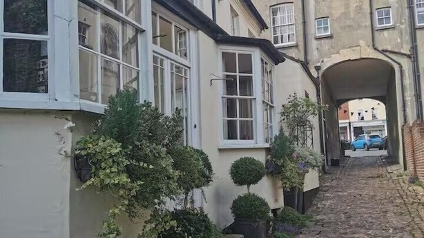 A narrow cobblestone street flanked by buildings with white-framed windows and small potted plants.
