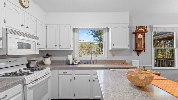 A kitchen with white cabinets, a stove, a microwave, a sink, and a window with a view of trees.