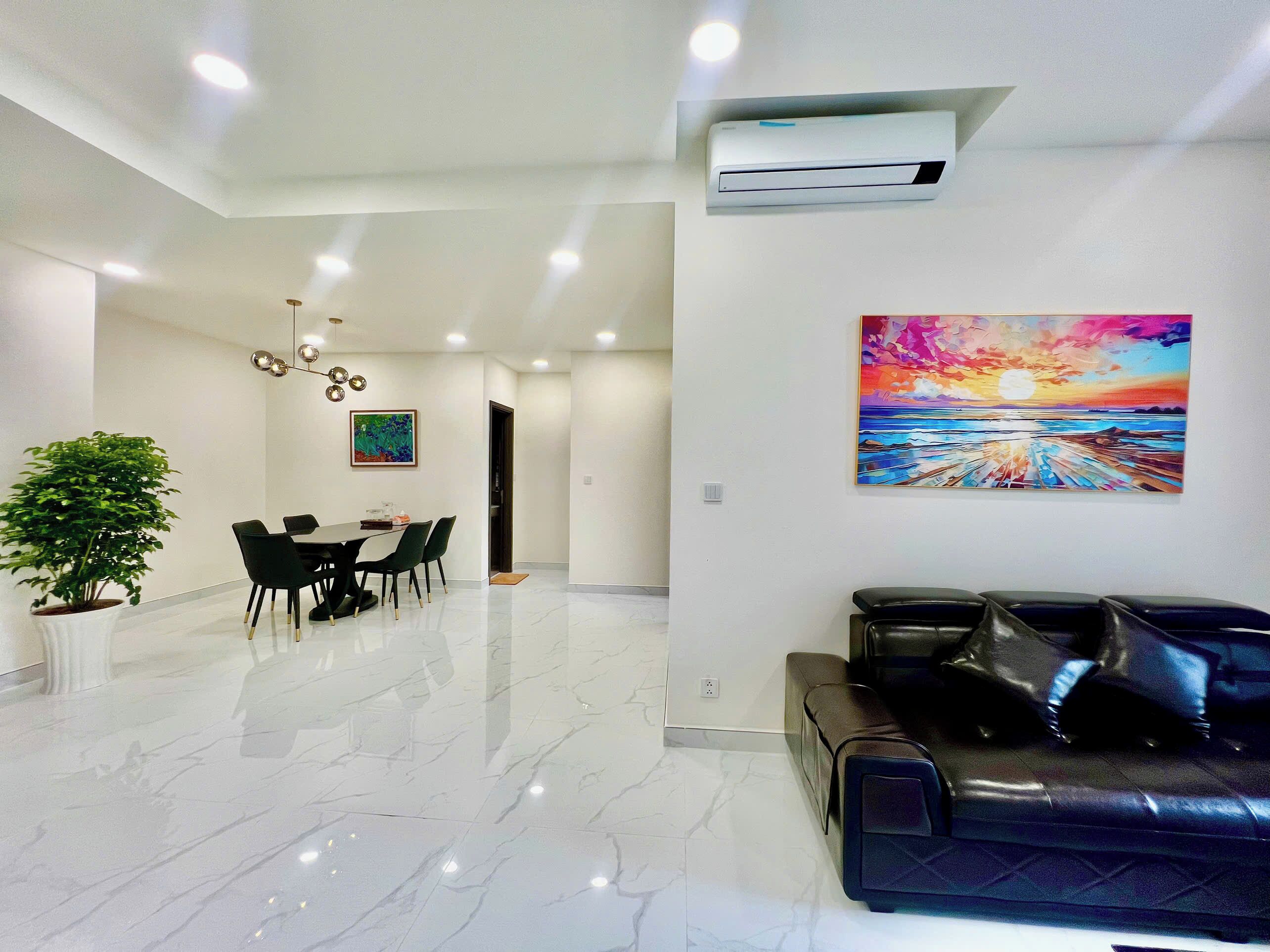 A modern living room with a black leather sofa, a dining area with a table and chairs, a large potted plant, and a vibrant sunset painting on the wall.