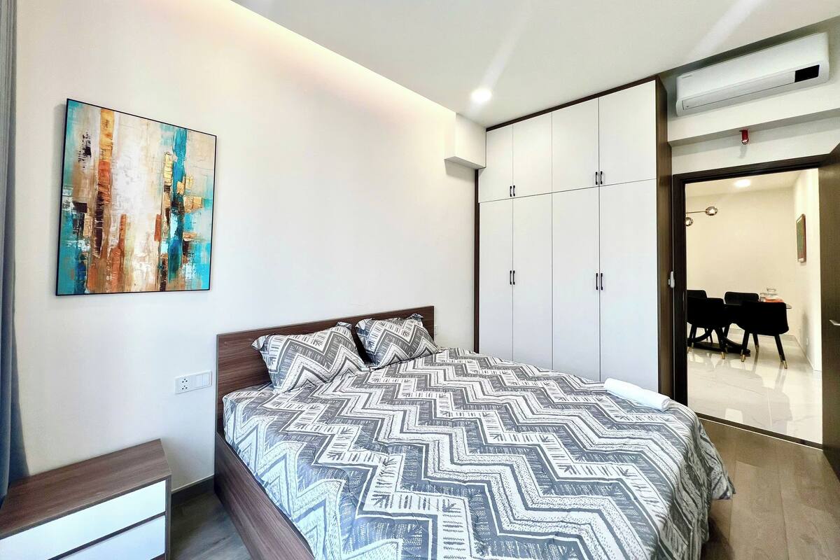 Une chambre à coucher avec un lit, une armoire, un tableau et un coin repas visible à travers une porte ouverte.