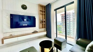 A modern living room with a flat-screen TV, a wooden entertainment unit, and a large window with a view of buildings. - CELEDON Tân Phú (Ho Chi Minh City)