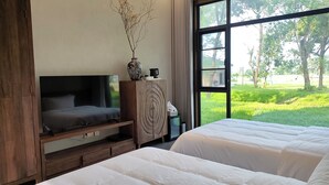 Un dormitorio con televisor, un armario de madera, un jarrón con ramas y vistas a un paisaje verde a través de amplios ventanales.