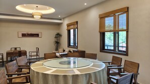 Un salón de conferencias con una mesa redonda de cristal, sillas de madera, una lámpara en el techo, una ventana con persianas de bambú y un cuadro en la pared.