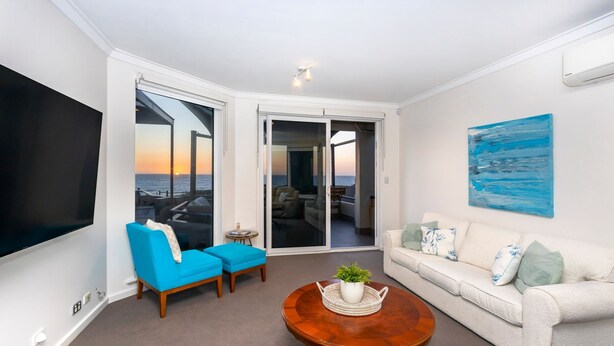 A living room with a large TV, two blue armchairs, a white sofa, and a coffee table with a plant. - Trigg Coastal House - Beachfront Bliss with Sunset Views (Trigg)