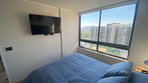 Un dormitorio con un ventanal grande, una cama con ropa de cama azul y un televisor montado en la pared.