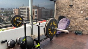 Un gimnasio con equipo de levantamiento de pesas, incluyendo barra y pesas, en un balcón con vista a la ciudad.