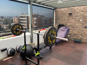 Et fitnessrum med styrketræningsudstyr, herunder vægtstang og vægte, på en balkon med udsigt over byen.