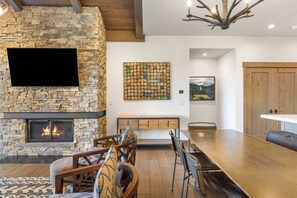 A modern living room with a stone fireplace, a flat-screen TV, a dining area with a long table, and a wooden ceiling. - Moonlight Basin Retreat | Ski-In/Out + Patio (Big Sky)