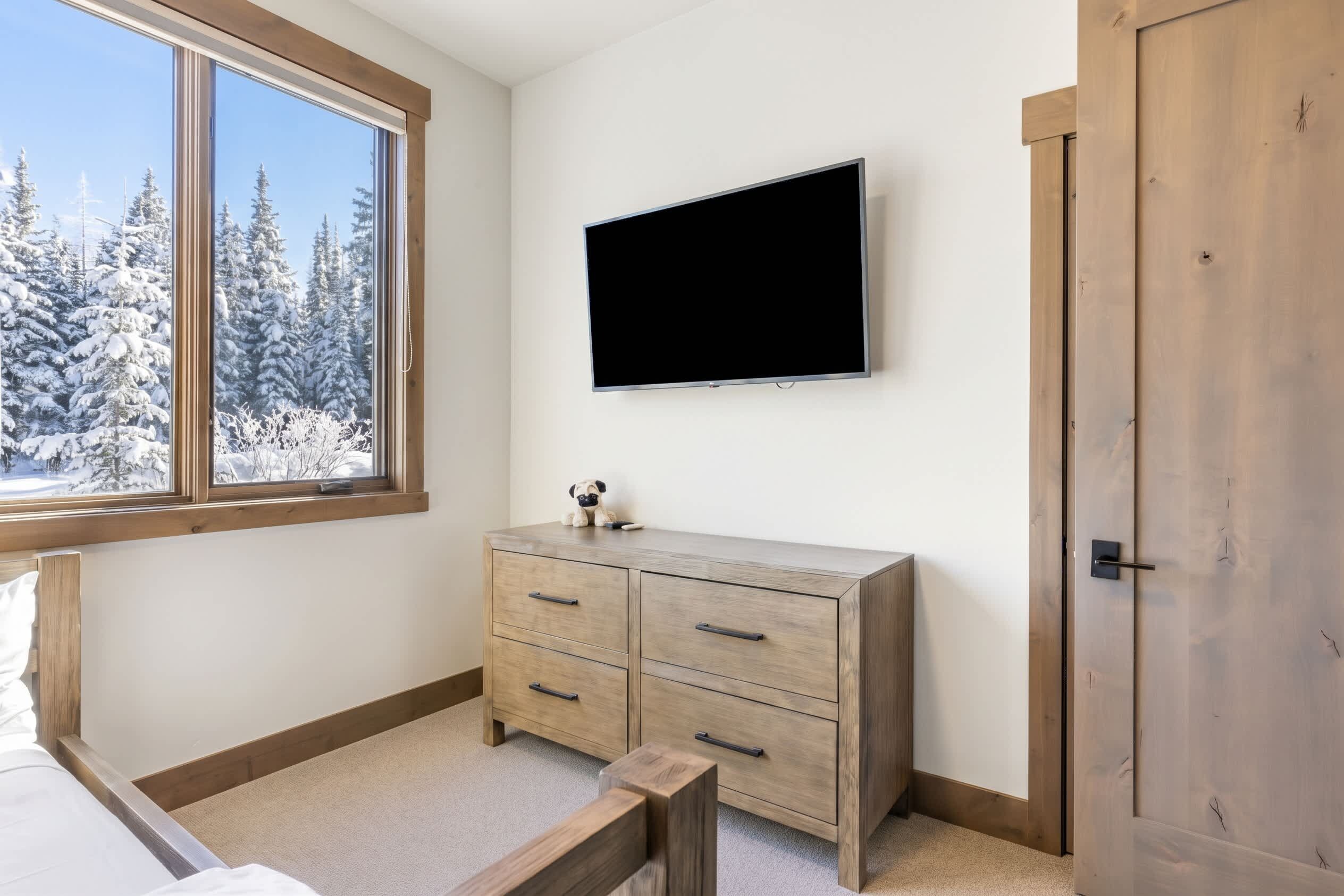 Een slaapkamer met een bed, een houten dressoir, een aan de muur bevestigde televisie en een raam met uitzicht op een besneeuwd bos.