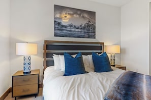 A hotel room with a bed, two bedside tables with lamps, a nightstand with a drawer, and a painting of a mountain landscape above the bed. - Moonlight Basin Retreat | Ski-In/Out + Patio (Big Sky)