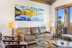 A living room with a leather sofa, two side tables with lamps, a coffee table, and a large painting of mountains and a lake. - Moonlight Basin Retreat | Ski-In/Out + Patio (Big Sky)