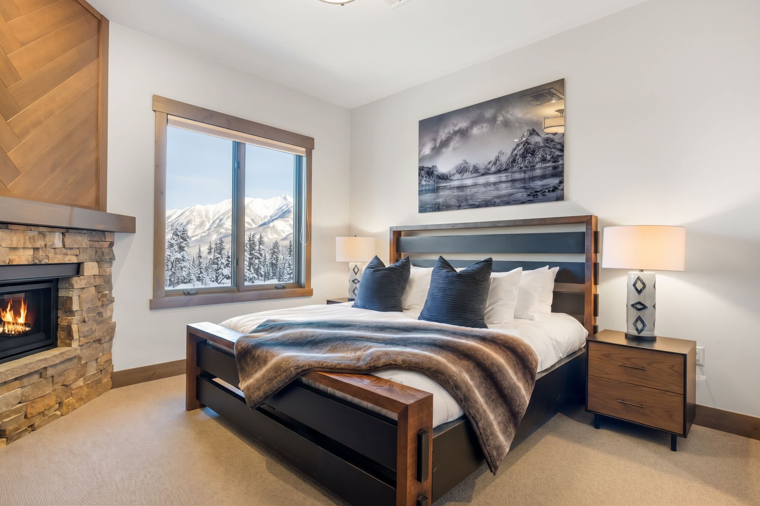 Een slaapkamer met een stenen openhaard, een groot bed met uitzicht op besneeuwde bergen, een schilderij van een berglandschap en een nachtkastje met een lamp.